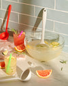 The Red Skinny Ladle on the far left corner and the Studio Ultimate Ladle in a bowl of juice with lemons and juice in glasses around it. 