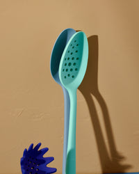The Teal Perforated spoon and Light Blue Ultimate Spoon, standing upright on a yellowish backgroun d